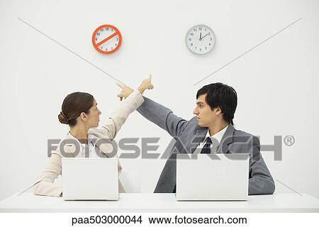 Picture - Professionals sitting at desk, pointing at clocks on wall behind them. Fotosearch