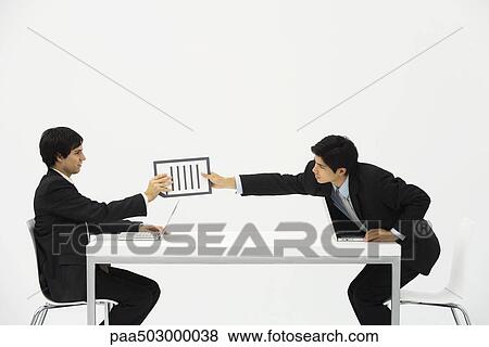 Two business associates sitting face to face at desk, one hand document to the other View Large Photo Image Stock Photo - Two business associates sitting face to face at desk, one hand document to the other. Fotosearch