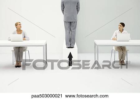 Stock Image - Two women sitting at desks, both looking at man standing above upward pointing arrow. Fotosearch