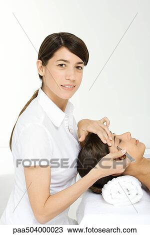 Stock Image - Beautician smiling at camera and plucking a woman's eyebrows. Fotosearch