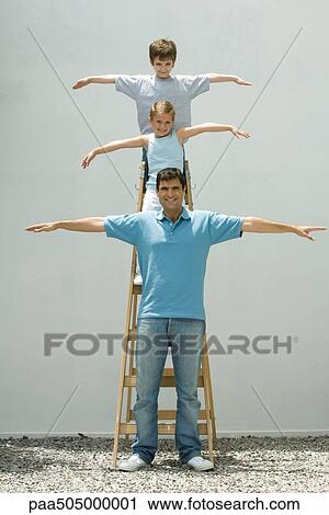 Stock Image - Boy and girl standing on ladder with arms in the air, father standing on the ground, all smiling at camera. Fotosearch
