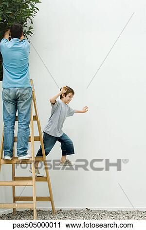 Stock Image - Father and son standing on ladder, boy leaping off. Fotosearch