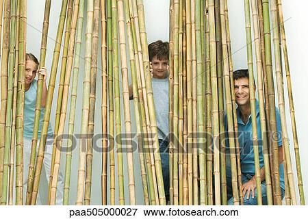 Stock Photo - Father and two children peeking through bamboo at camera, all smiling. Fotosearch