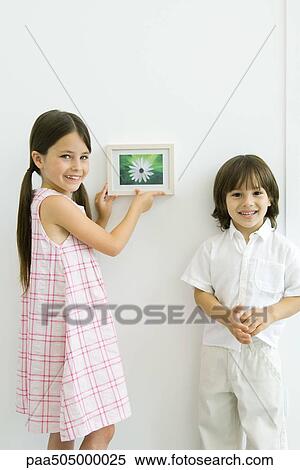 Stock Photography - Girl standing beside younger brother, hanging framed picture, both smiling at camera. Fotosearch
