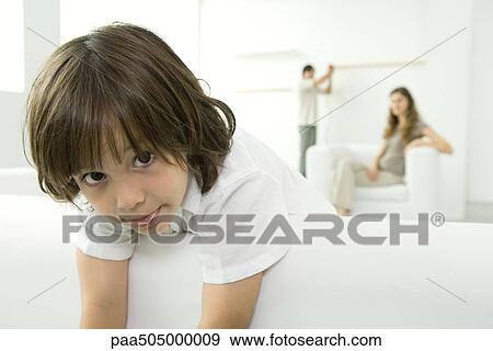 Stock Photo - Little boy leaning over couch, sticking tongue out at camera, parents in background. Fotosearch