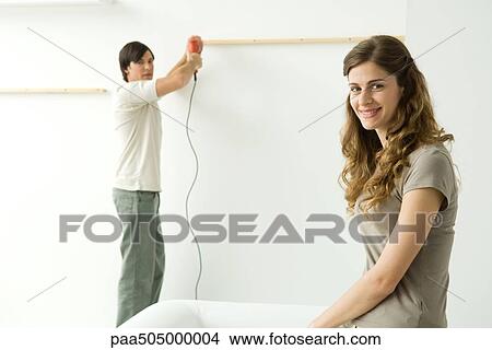 Man attaching thin piece of wood to wall, woman in foreground smiling at camera View Large Photo Image Picture - Man attaching thin piece of wood to wall, woman in foreground smiling at camera. Fotosearch