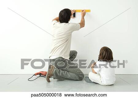 Stock Photo - Man measuring wall with a ruler while his son watches, drill on the floor nearby. Fotosearch