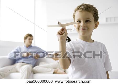 Stock Photograph - Boy playing with toy airplane, parents sitting on couch in background. Fotosearch