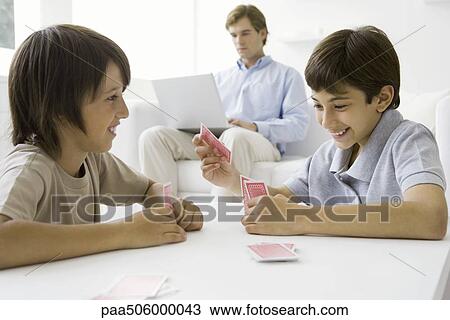 Stock Image - Brothers playing card game at table, father sitting on couch with laptop in background. Fotosearch