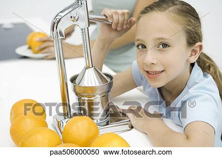 Stock Image - Little girl making fresh orange juice with citrus press, mother in background. Fotosearch