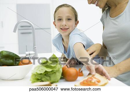 Stock Photo - Mother and daughter preparing sandwich together, girl smiling at camera, cropped view. Fotosearch