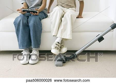 Two children sitting on sofa as parent vacuums, one playing handheld video game, cropped view View Large Photo Image Stock Photograph - Two children sitting on sofa as parent vacuums, one playing handheld video game, cropped view. Fotosearch