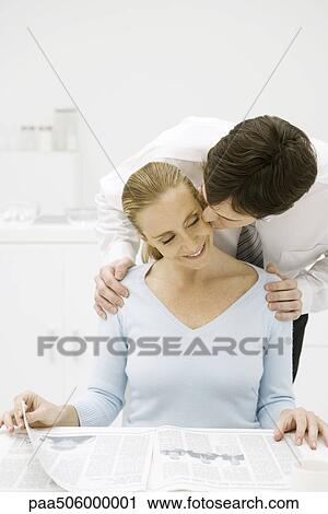 Stock Image - Woman sitting at table reading newspaper, husband leaning over her shoulder and kissing her on the cheek. Fotosearch