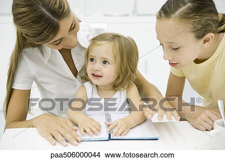 Picture - Woman sitting together with two daughters, looking at open notebook on table. Fotosearch