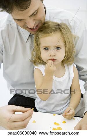 Young girl eating a piece of cereal, sitting on her father's lap, man helping her View Large Photo Image Stock Image - Young girl eating a piece of cereal, sitting on her father's lap, man helping her. Fotosearch