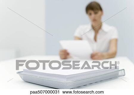 Stock Image - Stack of paper in metal tray, woman reading document in background. Fotosearch