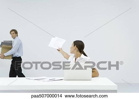 Woman sitting at desk, reaching back to hand documents to man carrying a box View Large Photo Image Picture - Woman sitting at desk, reaching back to hand documents to man carrying a box. Fotosearch