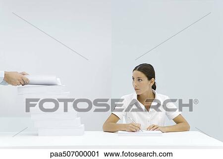 Stock Image - Woman sitting at table, looking at a staggered stack of paper, man's hands adding paper to the pile. Fotosearch