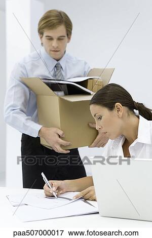Stock Photo - Woman writing on document at desk, man carrying cardboard box full of office supplies. Fotosearch