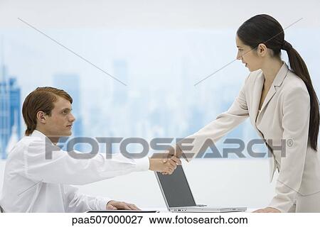 Young professionals shaking hands, man sitting, woman standing, side view View Large Photo Image Stock Photo - Young professionals shaking hands, man sitting, woman standing, side view. Fotosearch