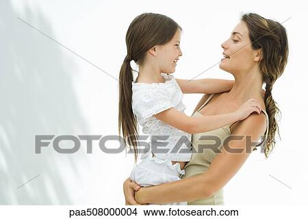 Picture - Mother holding and embracing daughter, both smiling at each other, side view. Fotosearch