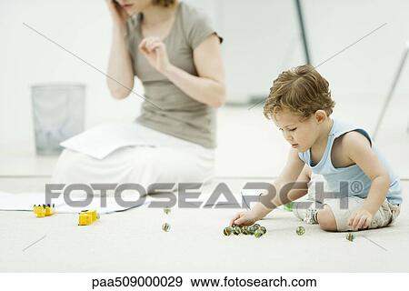 Little boy sitting on the ground, playing marbles, mother surrounded by paperwork in background View Large Photo Image Stock Photo - Little boy sitting on the ground, playing marbles, mother surrounded by paperwork in background. Fotosearch