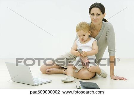 Stock Image - Professional woman sitting on the ground with laptop, holding toddler on lap, toddler looking at cell phone. Fotosearch