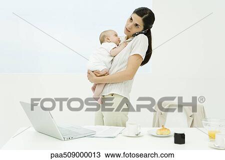 Stock Image - Professional woman standing at breakfast table, holding baby, using cell phone. Fotosearch
