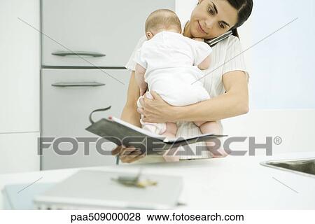 Woman holding baby, using cell phone and looking at agenda View Large Photo Image Stock Photo - Woman holding baby, using cell phone and looking at agenda. Fotosearch