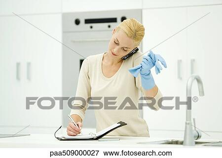 Woman standing at kitchen sink, wearing rubber gloves, using cell phone and writing in agenda View Large Photo Image Stock Photo - Woman standing at kitchen sink, wearing rubber gloves, using cell phone and writing in agenda. Fotosearch