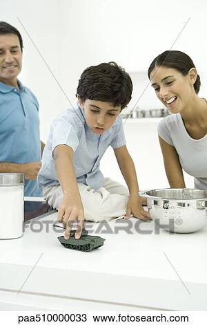 Stock Image - Boy playing with toy tank on kitchen counter, parents looking on and smiling. Fotosearch