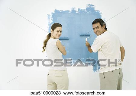 Stock Photo - Couple painting wall blue using rollers, both looking over their shoulders and smiling at camera. Fotosearch