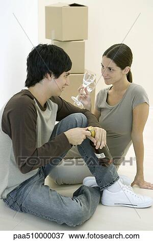 Stock Photo - Couple sitting on the floor together, woman holding pair of wine glasses, man opening wine bottle. Fotosearch