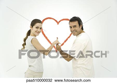 Stock Image - Couple standing in front of heart painted on wall, both holding onto the same paintbrush. Fotosearch