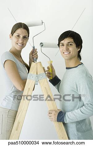 Picture - Couple standing on ladder, both holding up paint rollers and smiling at camera. Fotosearch