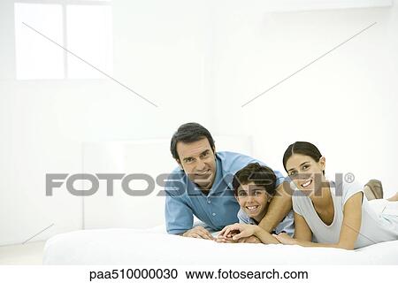 Stock Image - Family lying on bed together, holding hands, smiling at camera. Fotosearch