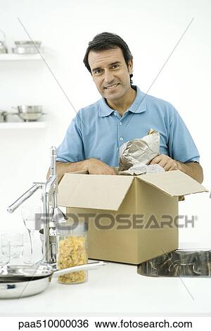 Stock Photograph - Man unpacking box on kitchen counter, smiling at camera. Fotosearch