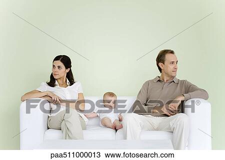 Stock Image - Couple sitting on couch with baby between them, both looking away. Fotosearch