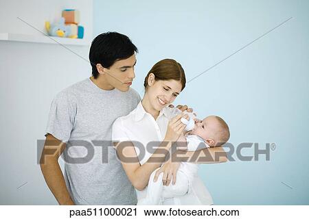 Stock Image - Mother feeding baby with a bottle, husband watching over her shoulder. Fotosearch