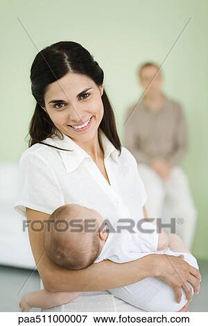 Stock Photo - Mother holding baby, smiling at camera, man in background. Fotosearch