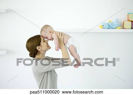 Stock Image - Mother lifting baby in the air, touching foreheads, side view. Fotosearch