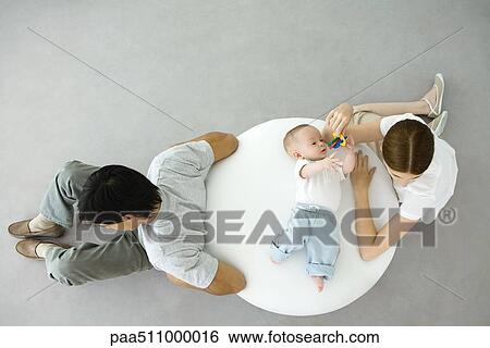 Mother playing with infant lying on ottoman, father sitting with back turned to them, overhead view View Large Photo Image Stock Photograph - Mother playing with infant lying on ottoman, father sitting with back turned to them, overhead view. Fotosearch
