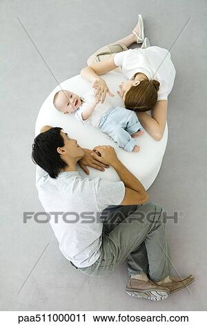 Stock Image - Parents and baby relaxing on ottoman, baby holding mother's hand, overhead view. Fotosearch