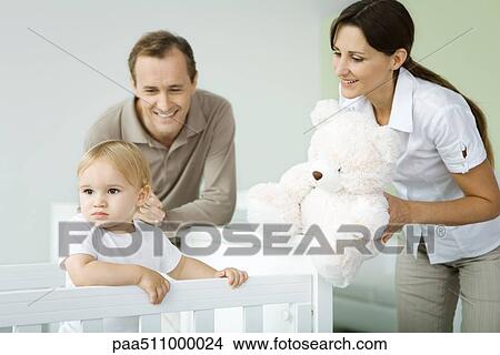 Picture - Parents standing beside toddler in crib, mother holding teddy bear, toddler looking away. Fotosearch