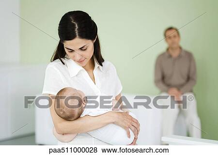 Stock Photo - Woman holding sleeping baby, husband sitting in background. Fotosearch