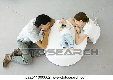 Stock Image - Young parents leaning against ottoman, looking at baby lying between them. Fotosearch