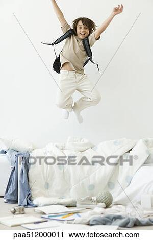 Stock Image - Boy jumping on bed in messy room, arms raised. Fotosearch