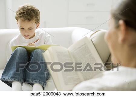 Boy sitting on sofa, writing in book, mother reading in foreground View Large Photo Image Stock Photo - Boy sitting on sofa, writing in book, mother reading in foreground. Fotosearch