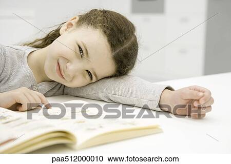 Girl sitting at table with book, resting head on arm, smiling at camera View Large Photo Image Stock Image - Girl sitting at table with book, resting head on arm, smiling at camera. Fotosearch