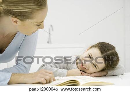Stock Photography - Mother and daughter looking at book together, girl resting head on arms. Fotosearch